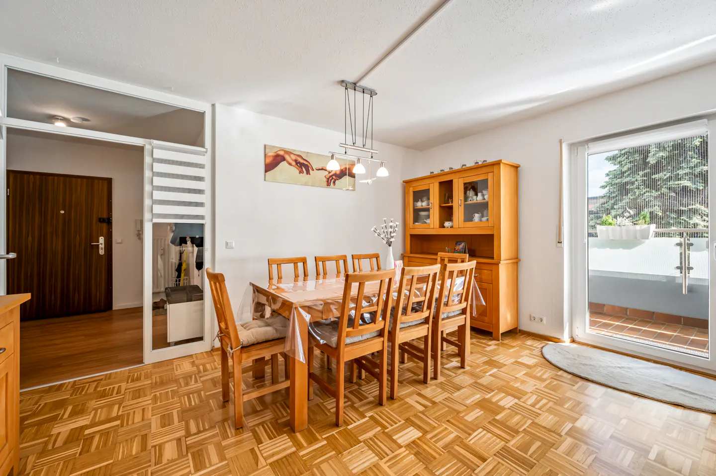 Bright dining room with wood parquet floor, table with chairs, hutch, and balcony door. A painting hangs on the white wall.