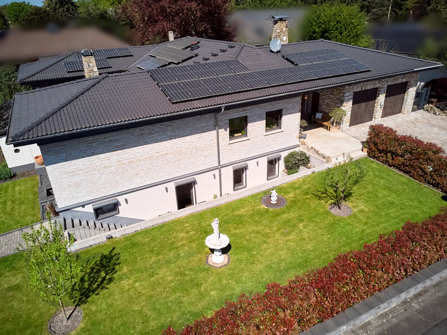 Aerial view of a two-story house with a dark roof, solar panels, stone facade, green lawn, and a fountain.
