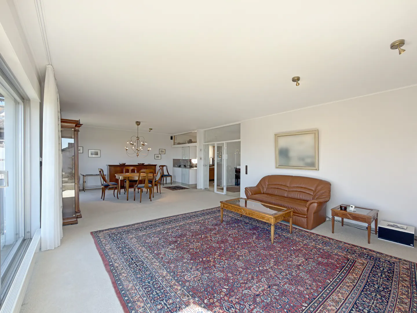A bright, open-concept living and dining area with a brown leather sofa, a large patterned rug, and a dining table.