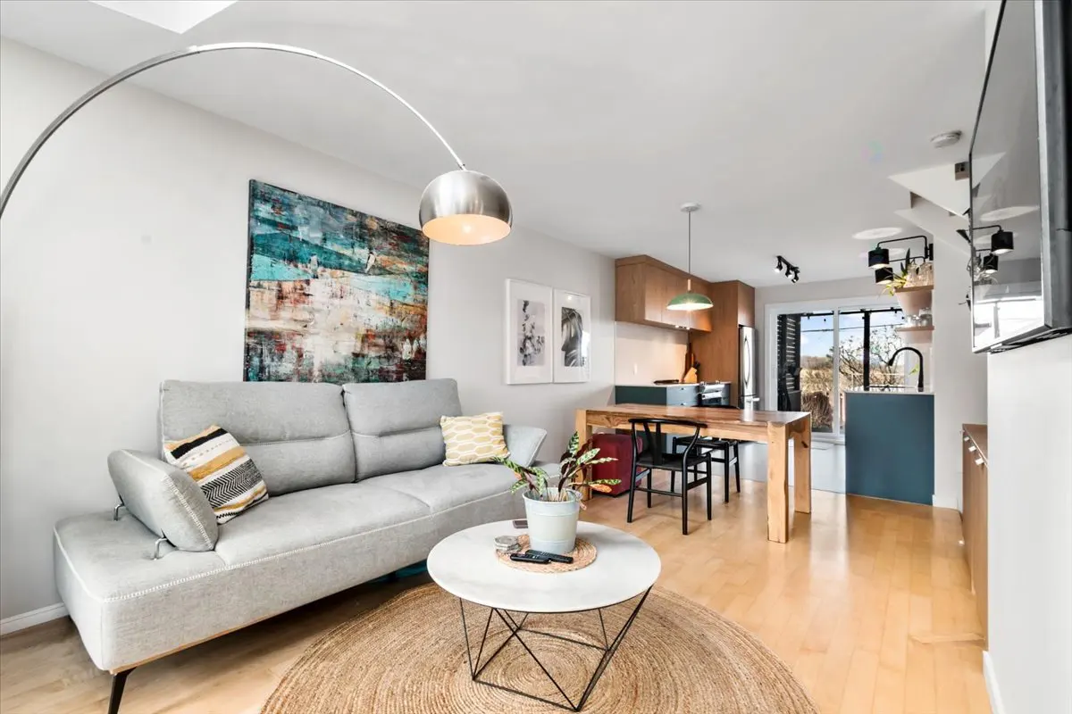 Bright, modern living room with a gray sofa, round jute rug, and a white marble coffee table. An open kitchen is visible in the background.