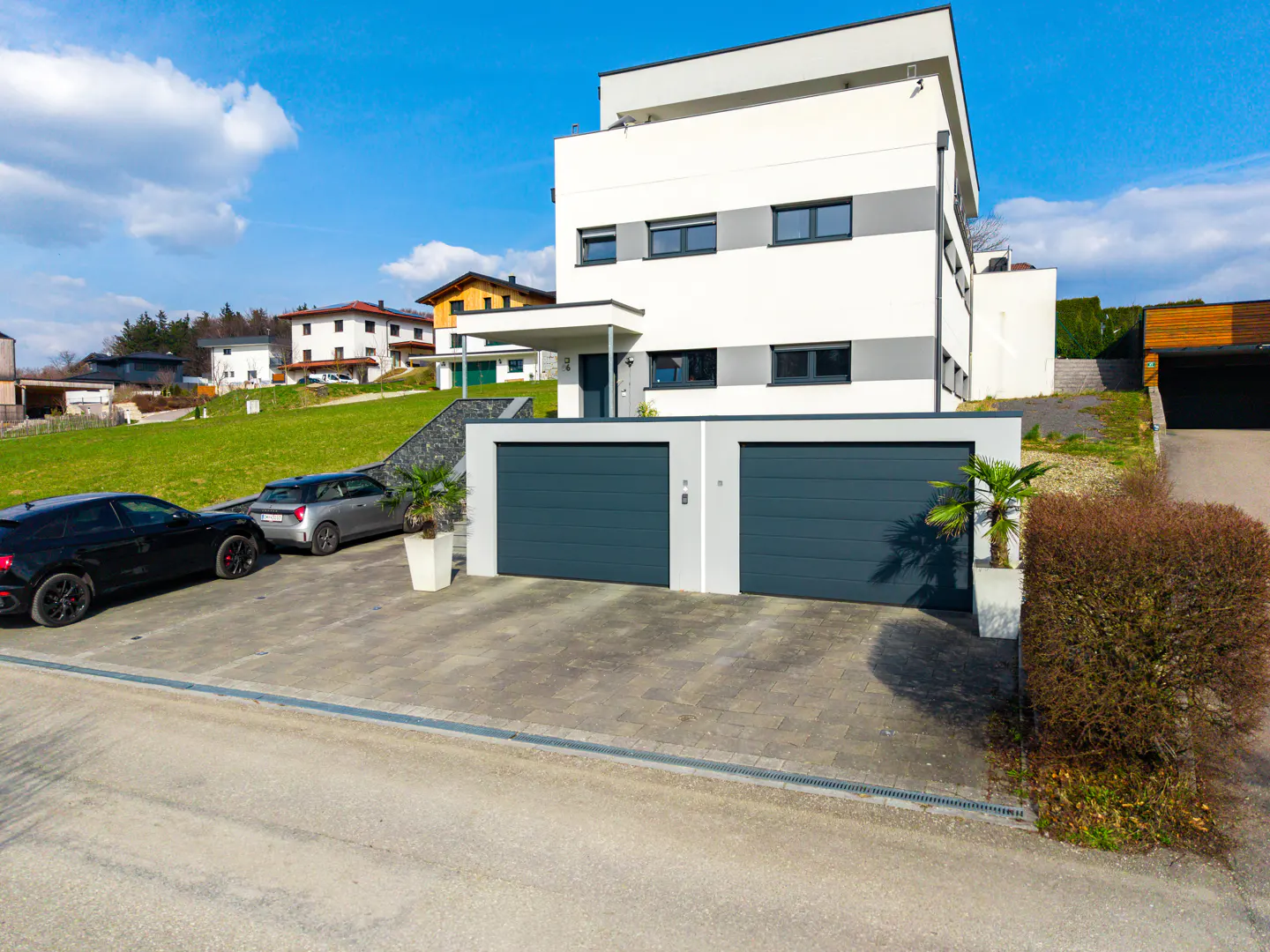 Modern white house with gray accents, two-car garage, and driveway with parked cars under a blue sky.
