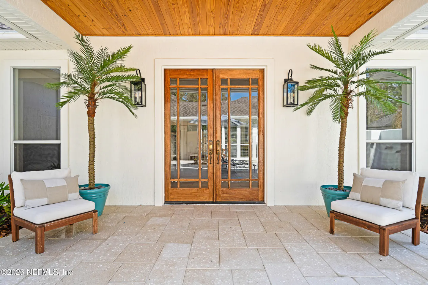 Exterior view of a home featuring double wood doors, two chairs, and two potted palm trees.
