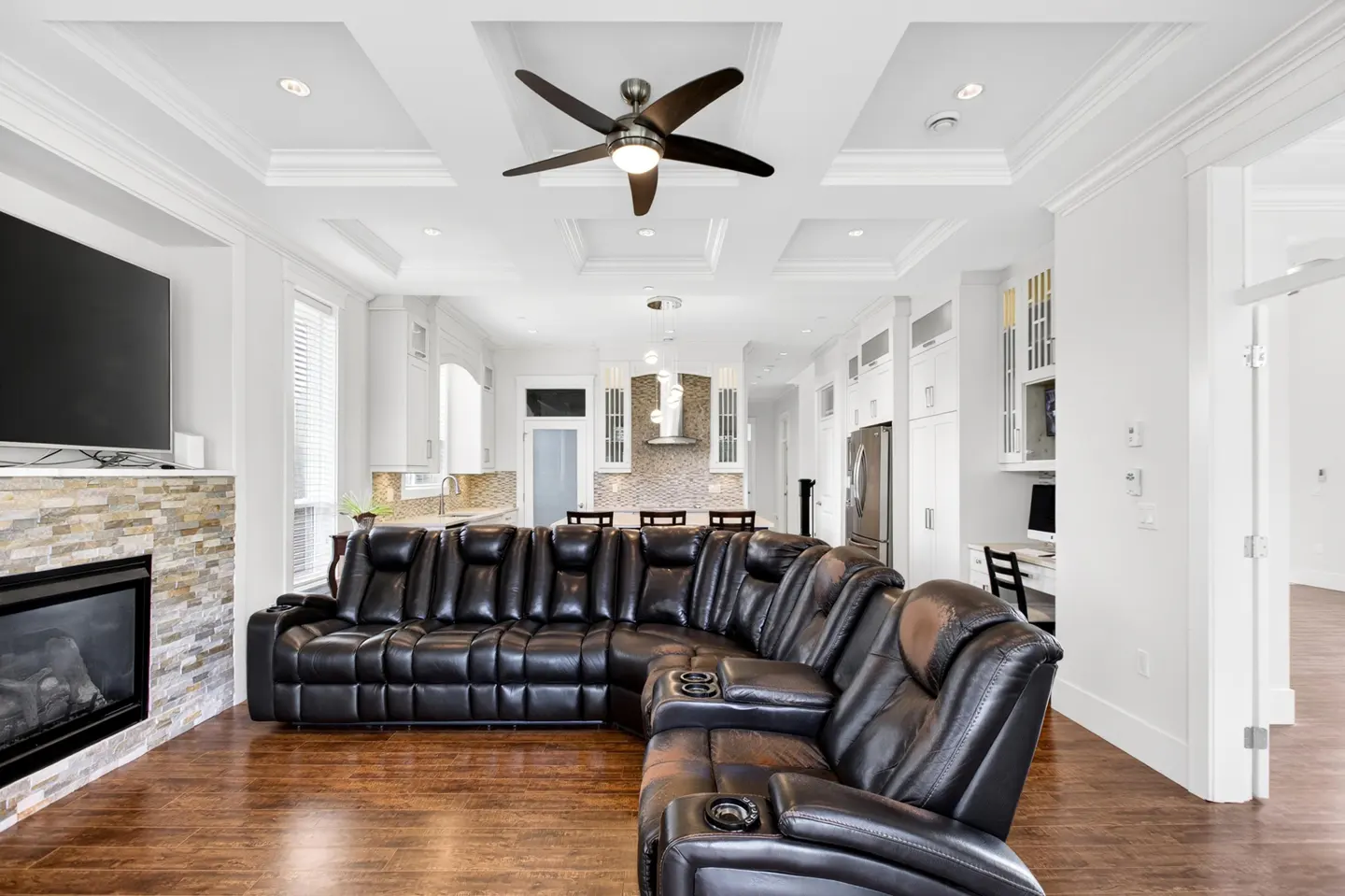 Bright, open-concept living room with a large black leather sectional sofa, fireplace, and kitchen in the background. White walls and ceiling.