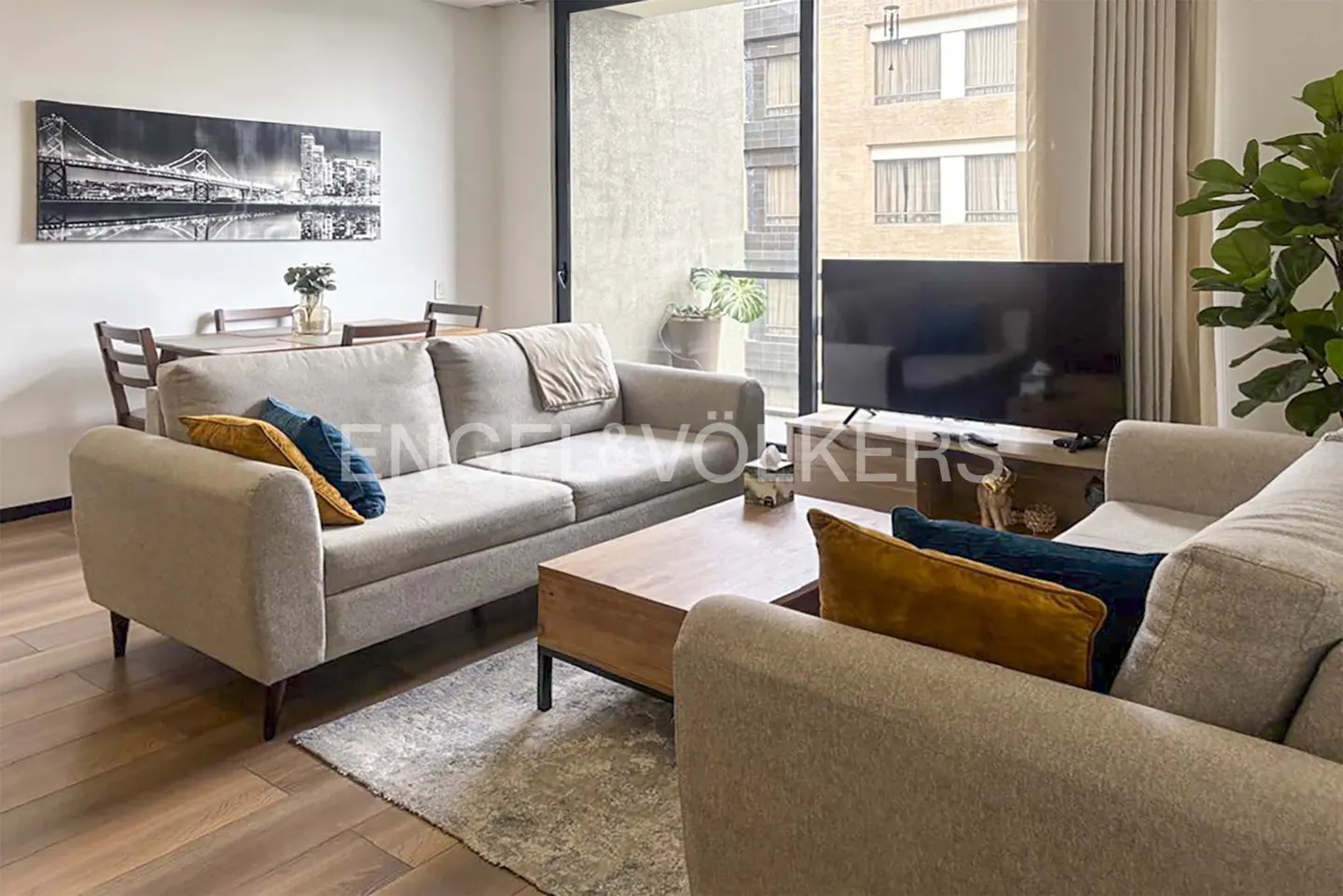 Living room with gray sofas, wood coffee table, and TV. A black and white bridge art hangs above a dining table. Balcony doors let in natural light.