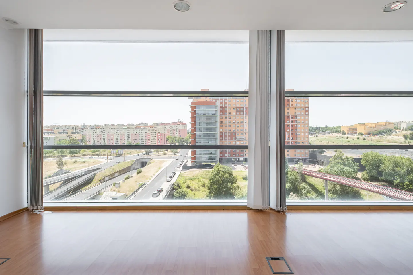 Empty room with wood floors and a large window showing a city view with buildings, roads, and greenery.