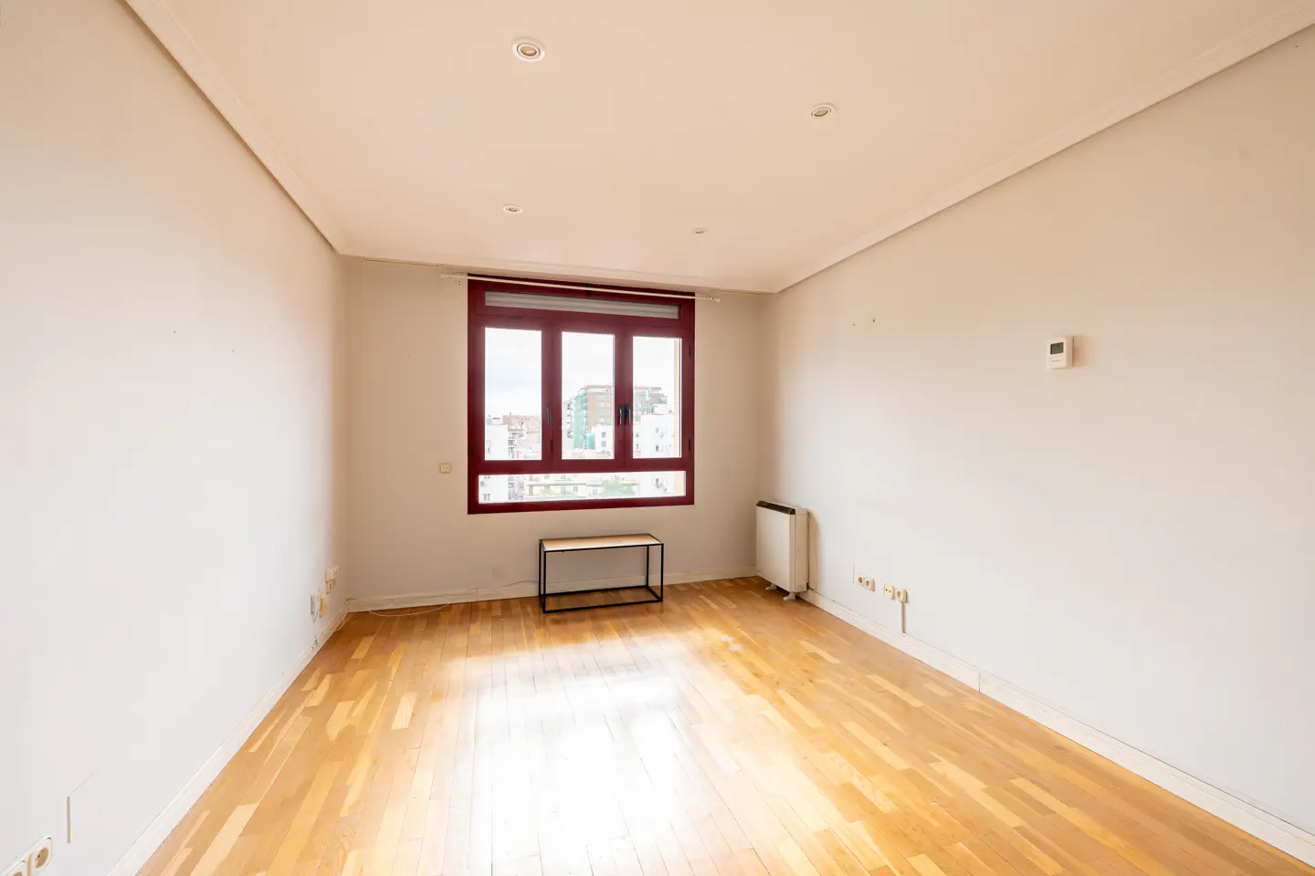 Empty room with light wood floors, white walls, and a red-framed window. A small table sits beneath the window.