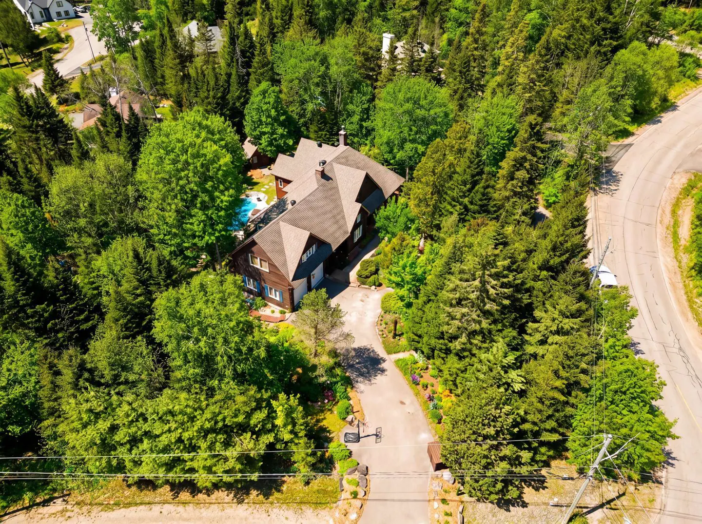 Aerial view of a brown house with a gray roof surrounded by green trees, a pool, and a driveway with a basketball hoop.