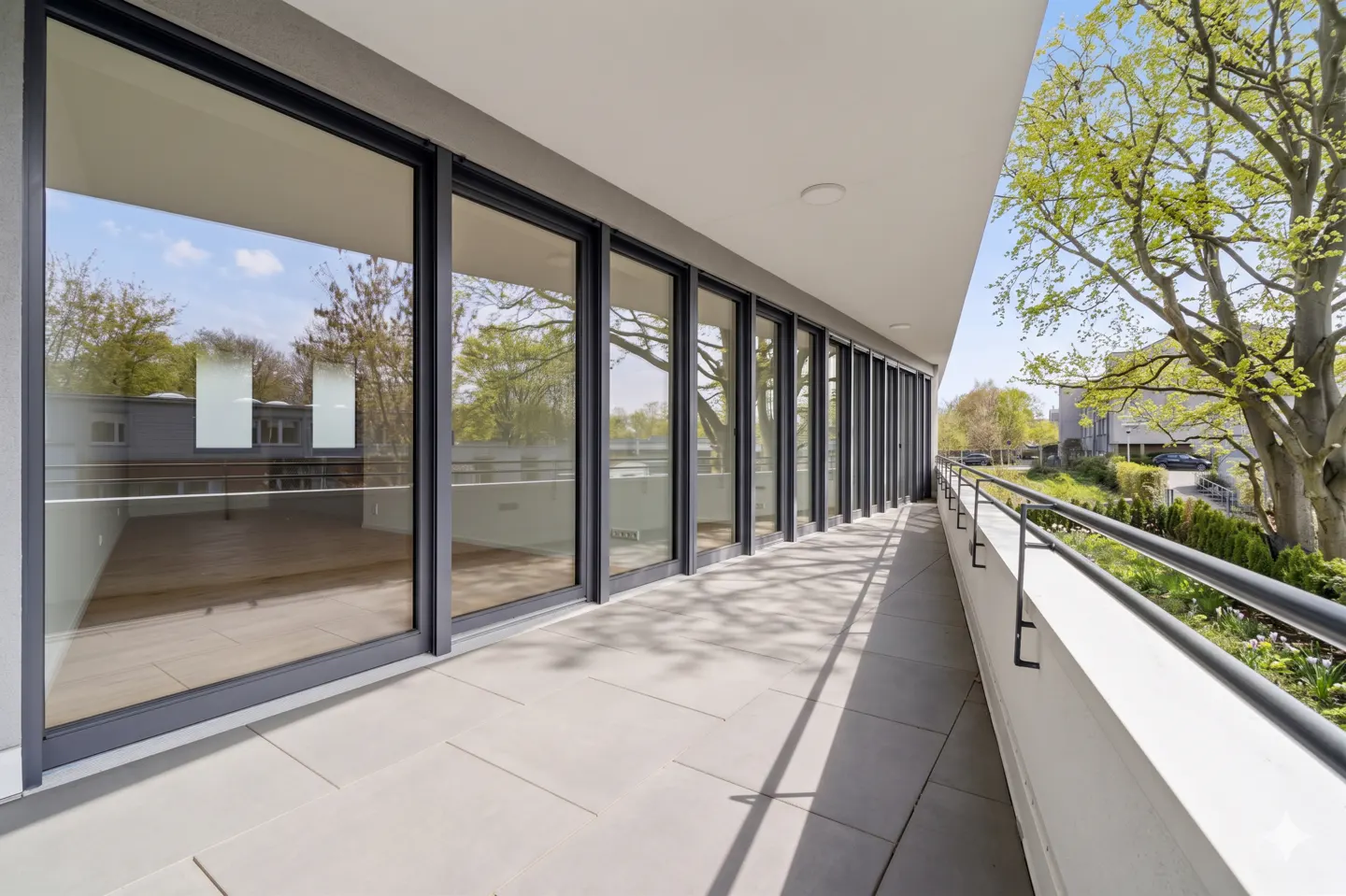 A long, gray-tiled balcony with black railings and floor-to-ceiling windows reflecting trees and sky.