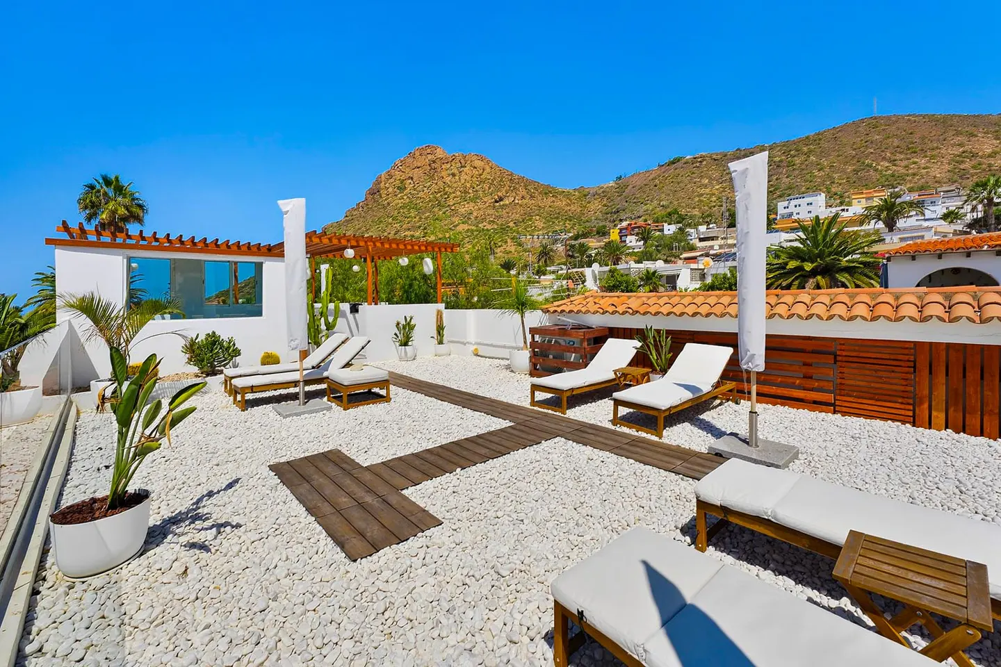 Sunny rooftop terrace with white gravel, wooden walkways, lounge chairs, umbrellas, and mountain views under a blue sky.
