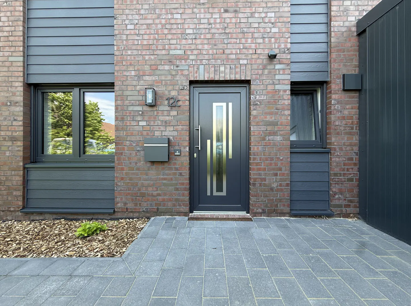 Modern home exterior with brick and gray siding. A gray door with gold accents is centered, with a mailbox and window to the left. Paved walkway.