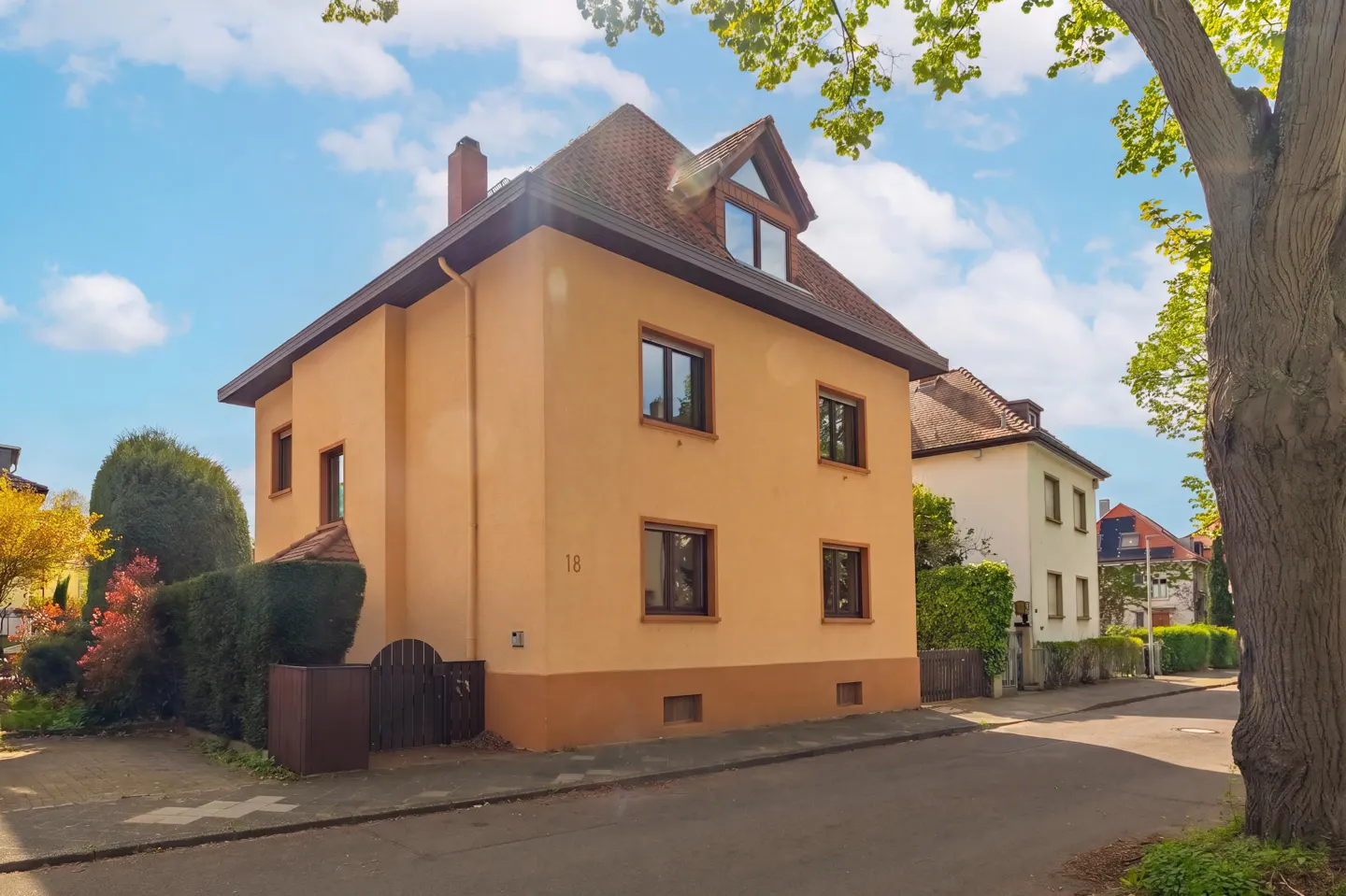Two-story peach house with brown trim and a red tile roof on a tree-lined street under a blue sky.