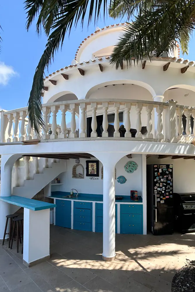 Outdoor kitchen with blue cabinets, white walls, and a black refrigerator. Palm tree fronds are visible in the foreground.