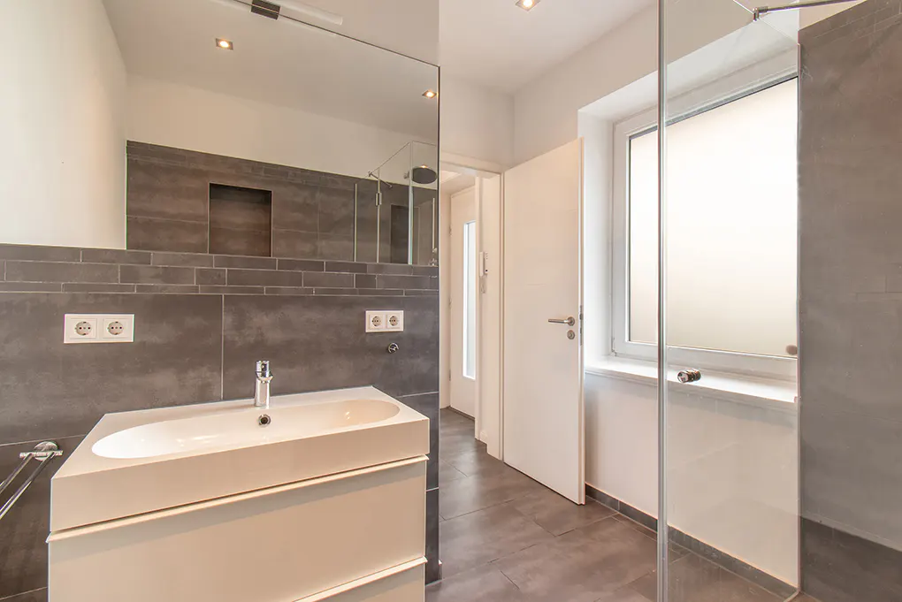 Modern bathroom with gray tile, white sink vanity, and glass shower enclosure. A white door is partially open.