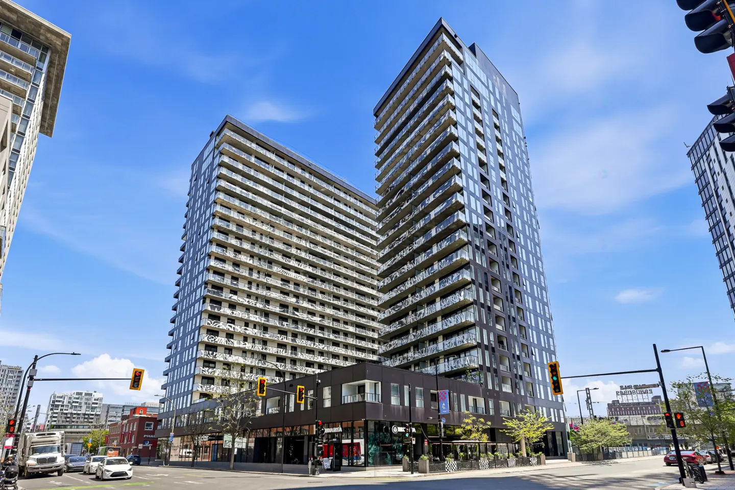 Two modern high-rise apartment buildings with glass balconies under a blue sky. Street-level retail and traffic lights are visible.