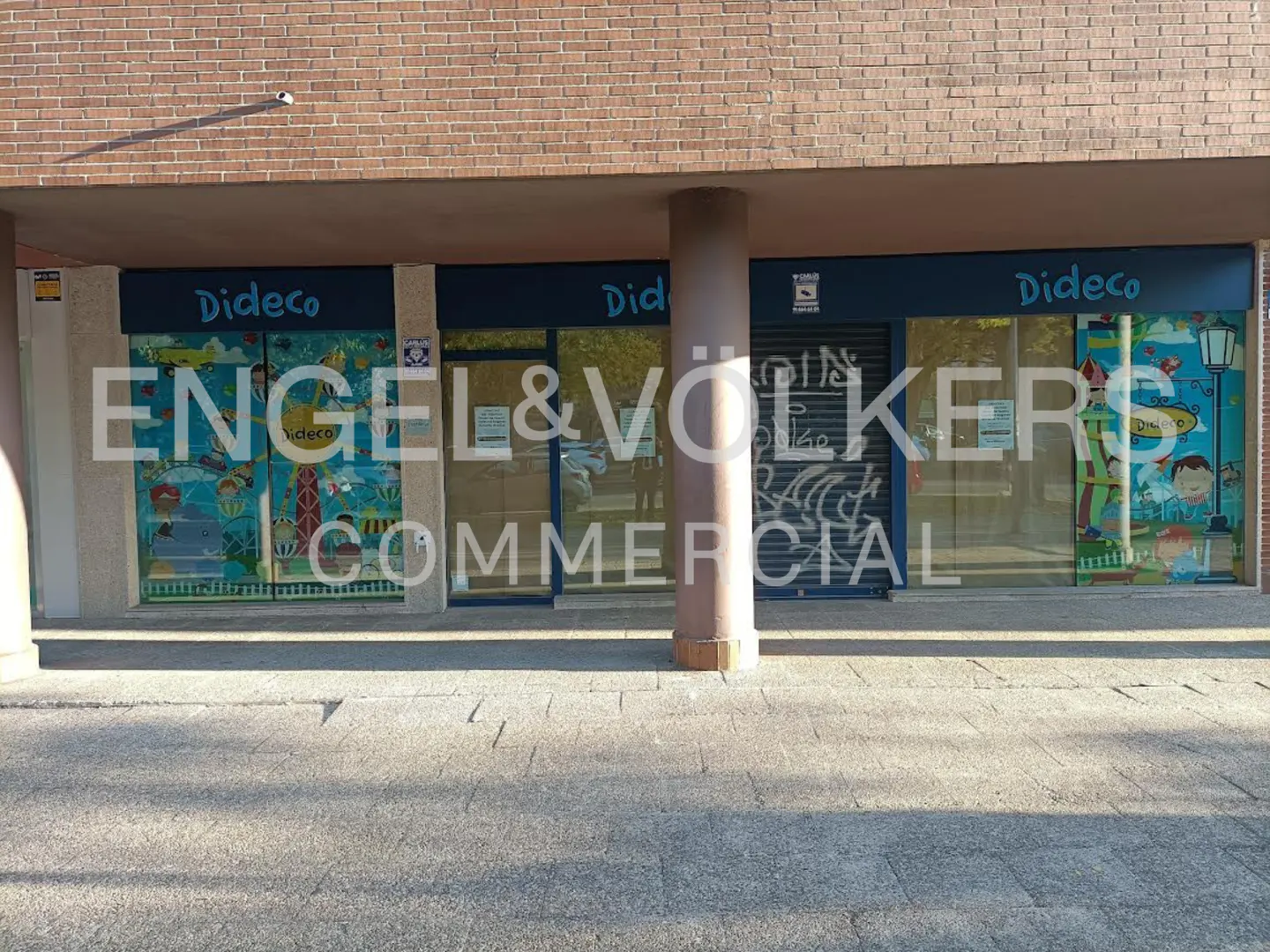 Storefront of "Dideco" with colorful window art, under a brick building overhang. One window has graffiti and a closed shutter.