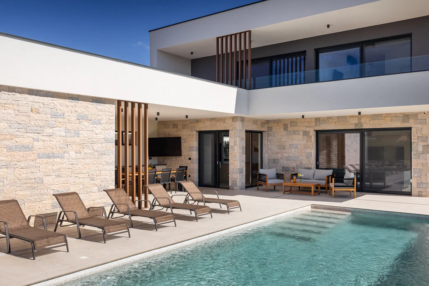 Modern luxury villa with a stone facade, swimming pool, and patio featuring lounge chairs and outdoor dining area.