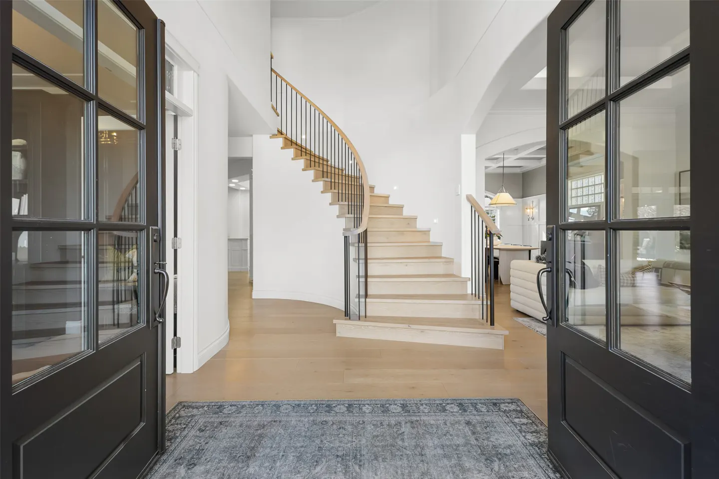 Open black double doors reveal a foyer with a curved staircase, light wood floors, and a blue patterned rug.
