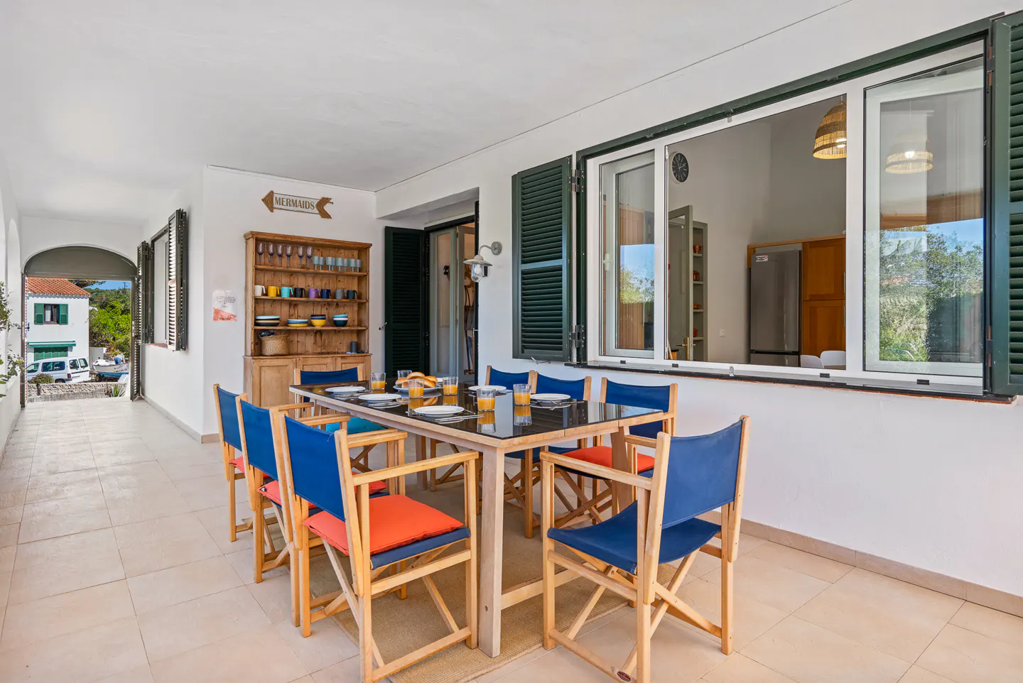 Outdoor dining area with a black table, blue and orange chairs, and a wooden hutch. A window looks into the kitchen.