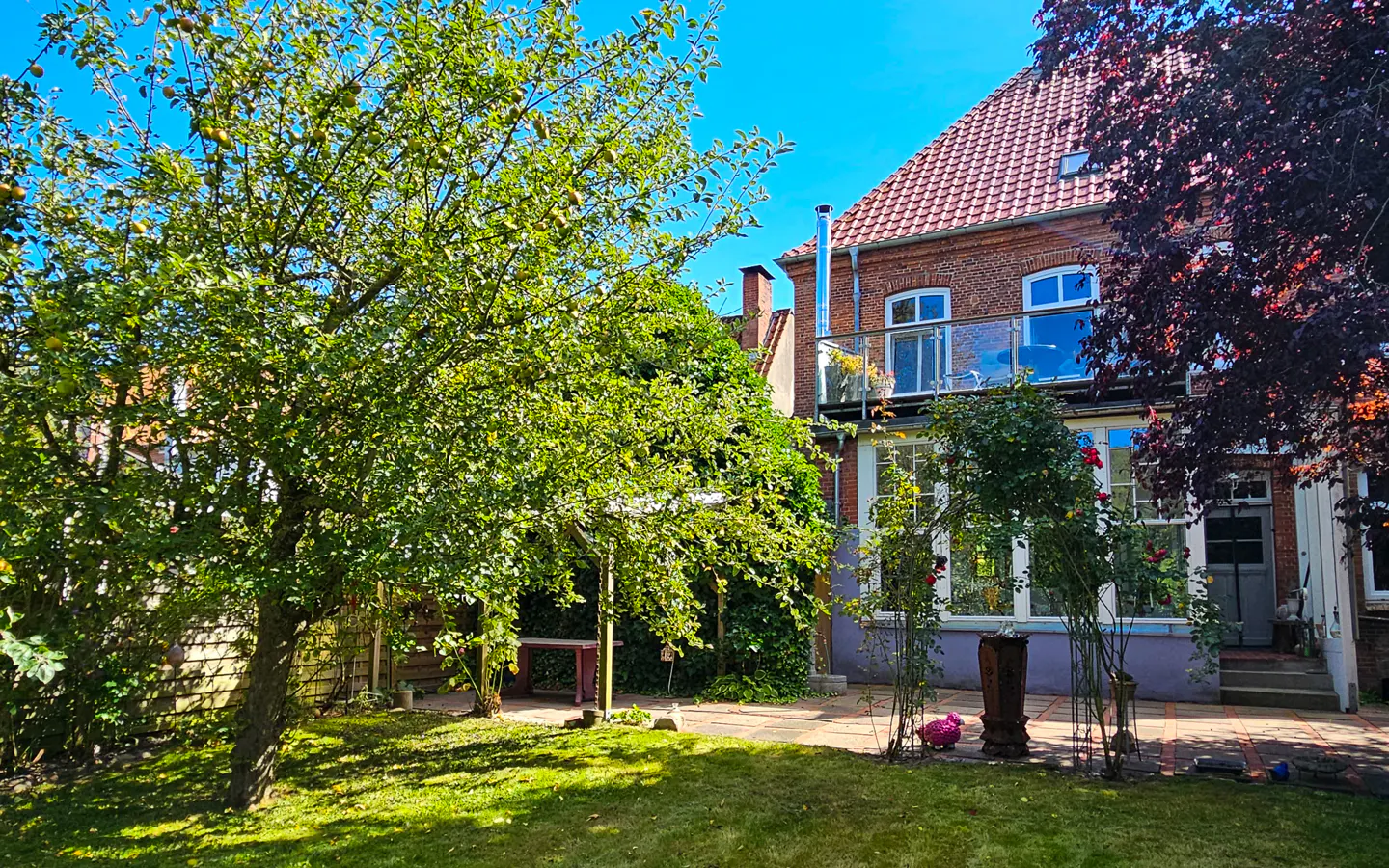 Brick house with a red tile roof, a glass balcony, and a lush green garden with trees and flowers.