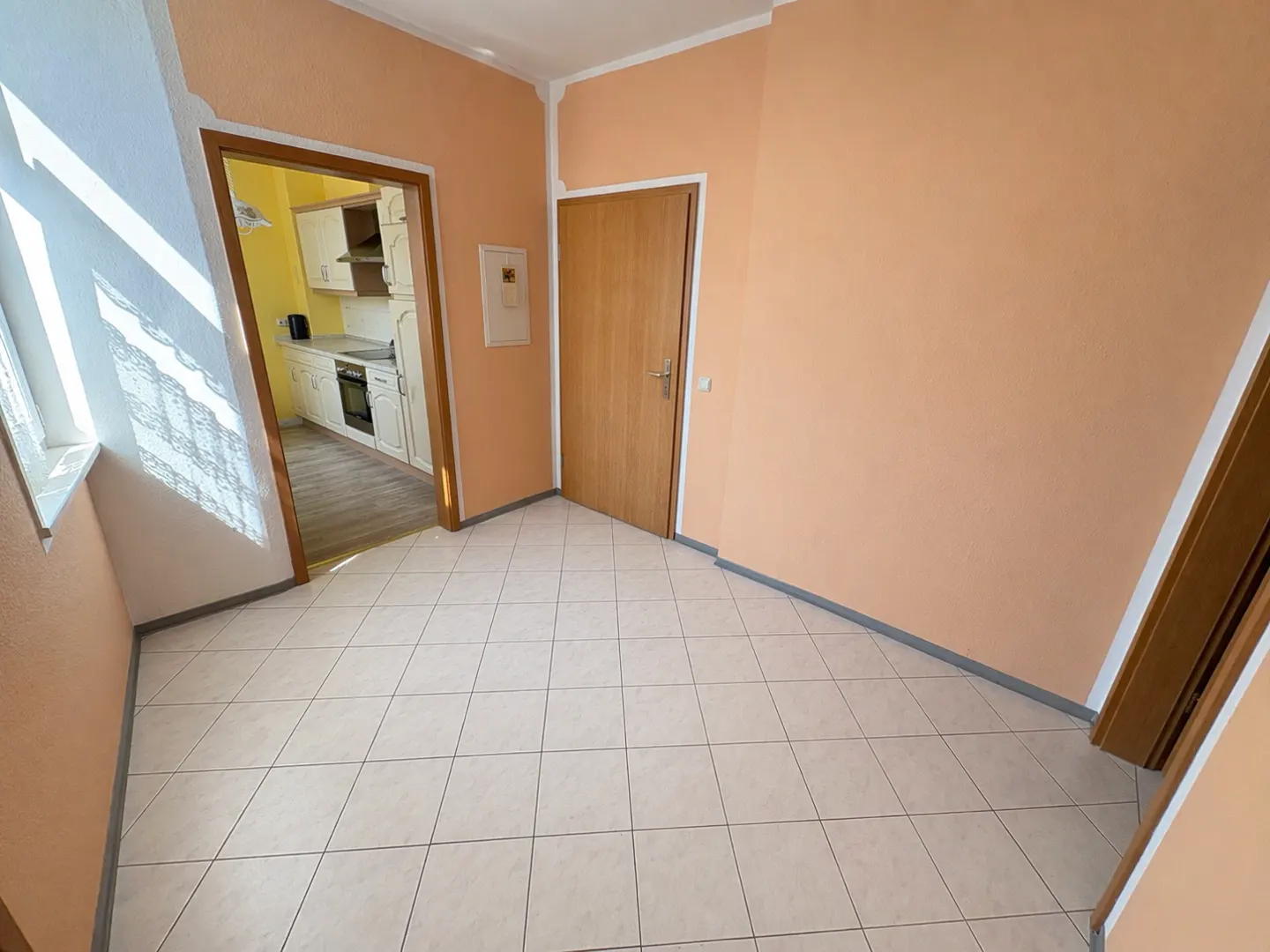 A room with peach walls and white tile floors. A doorway leads to a yellow kitchen with white cabinets. A closed wooden door is on the right.