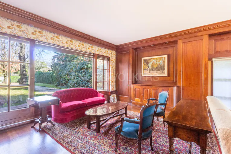 Living room with wood paneling, a red sofa, blue chairs, and a patterned rug. A large window overlooks a green garden.