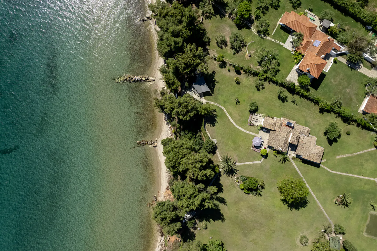 Aerial view of two waterfront villas with terracotta roofs, lush green lawns, and trees along a sandy beach and blue sea.