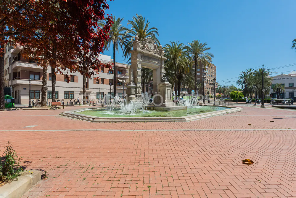 A sunny plaza with a stone fountain, palm trees, and brick paving. Buildings line the background, with a "Policia Local" sign visible.