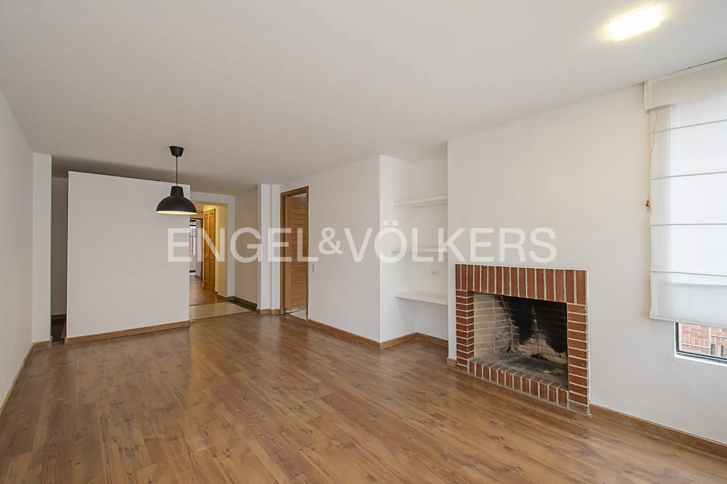 Bright living room with wood floors, white walls, and a brick fireplace. A black pendant light hangs from the ceiling. Engel & Völkers logo is visible.