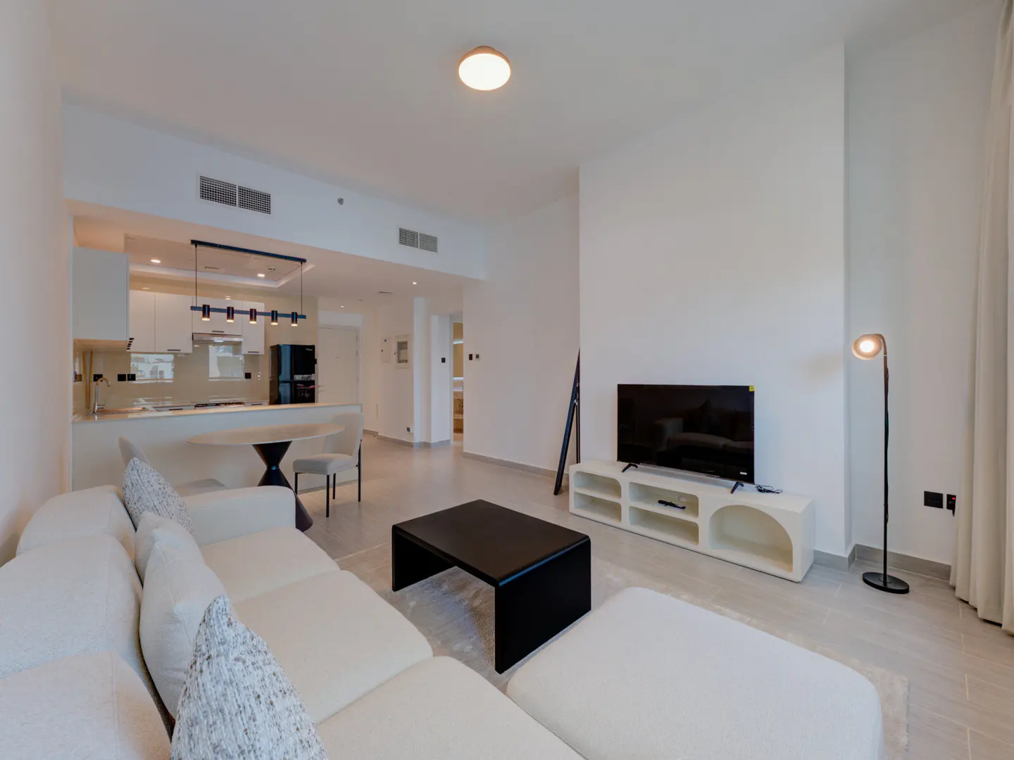 Bright, modern apartment living room with white sectional sofa, black coffee table, TV, and open kitchen in the background.
