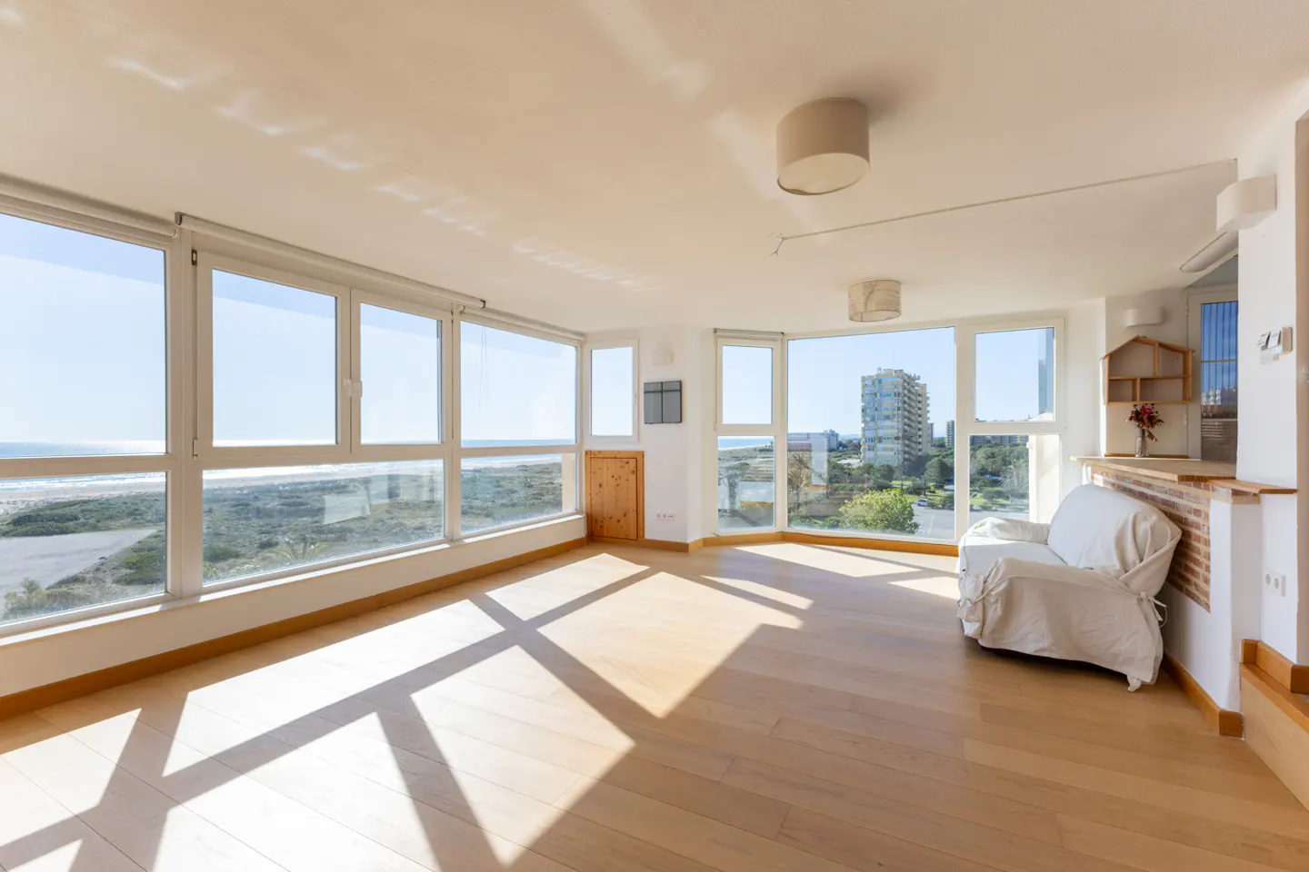 Bright, empty living room with wood floors and large windows overlooking the ocean. A white armchair sits near a brick half-wall.