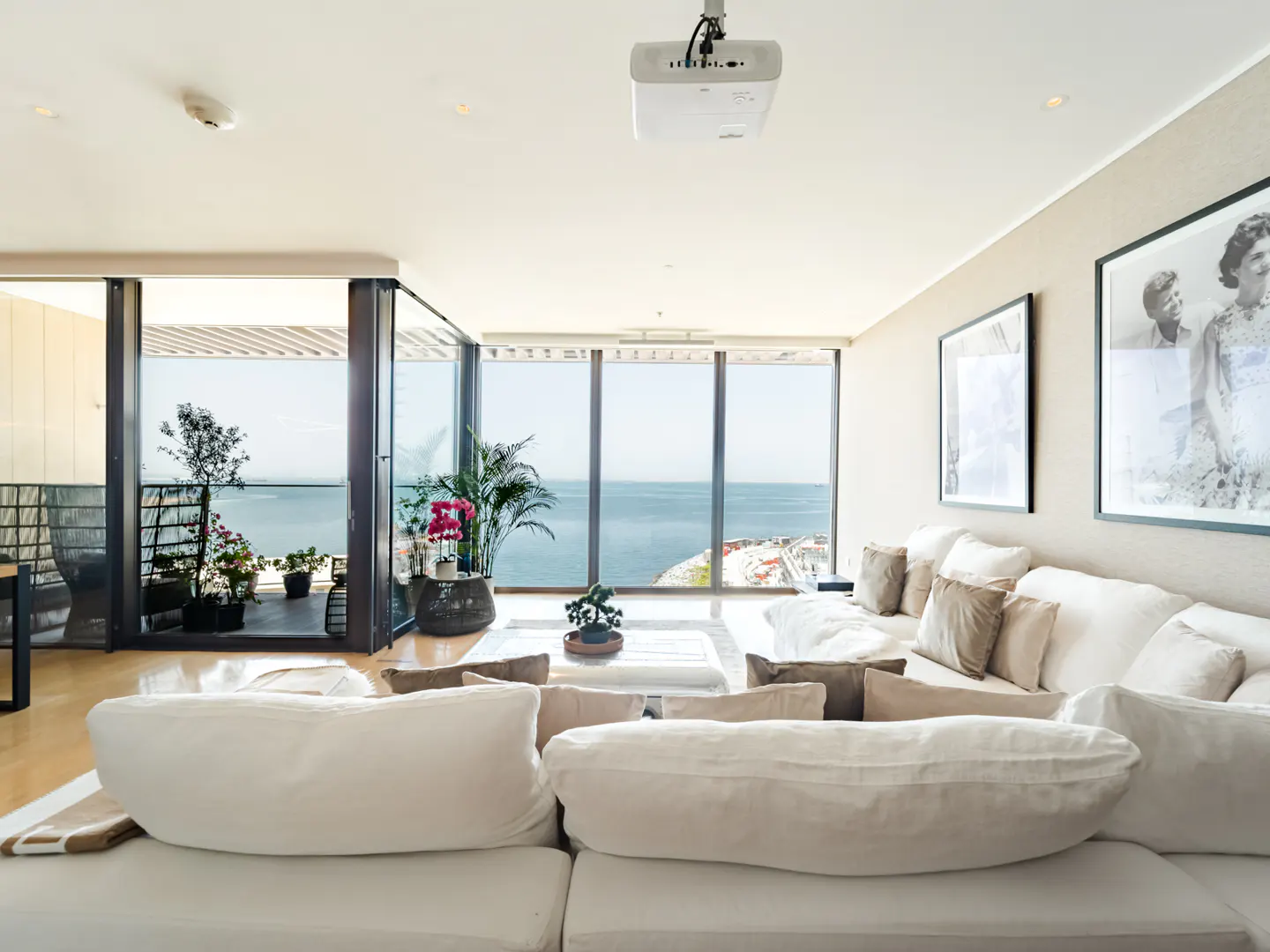 Bright living room with white sofa, beige pillows, and ocean view through large windows. A projector hangs from the ceiling.