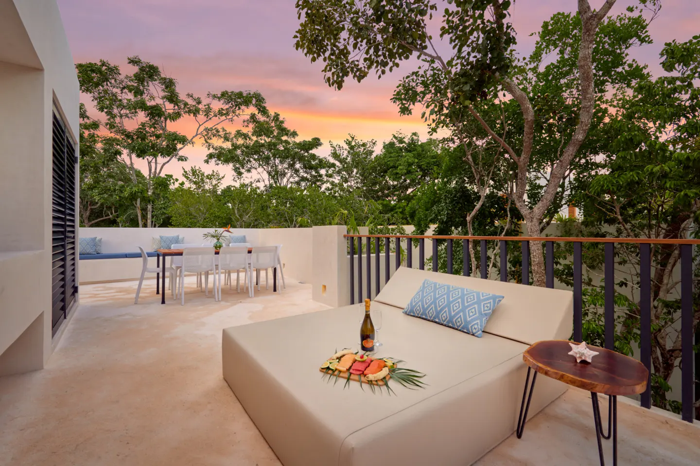 Outdoor terrace with a lounge bed, fruit platter, and dining area overlooking lush trees at sunset.
