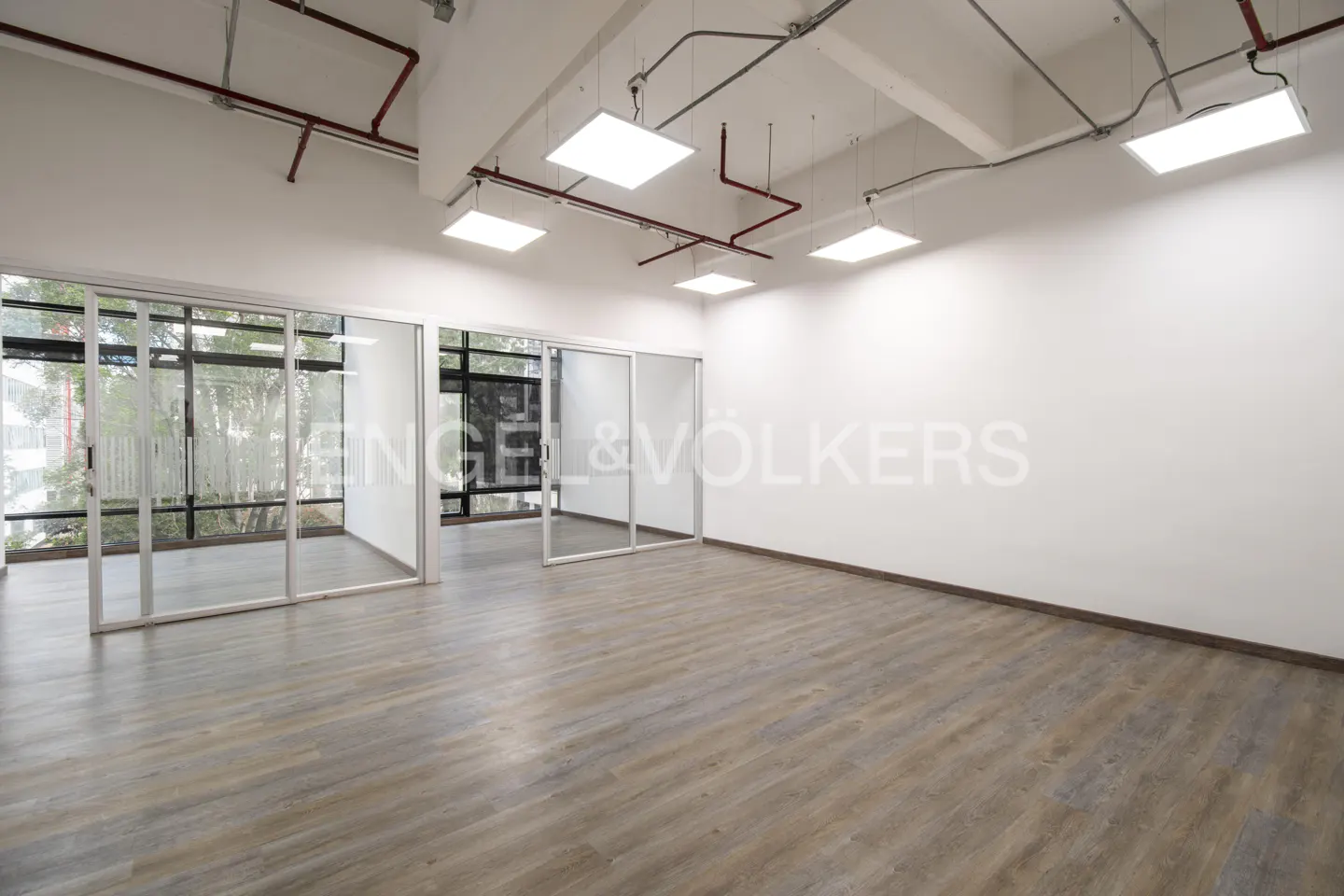 Empty office space with wood floors, white walls, and glass-walled rooms. Red pipes and square lights hang from the ceiling.