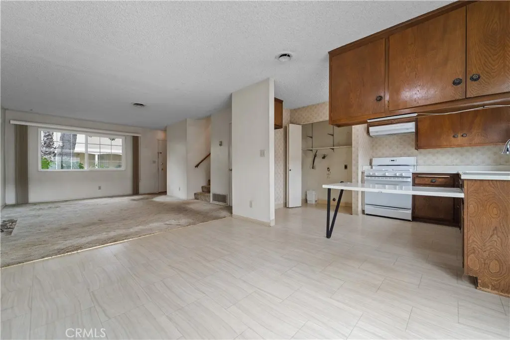 A bright, empty home interior with wood cabinets, white appliances, and tile flooring. A living room with a large window is visible.