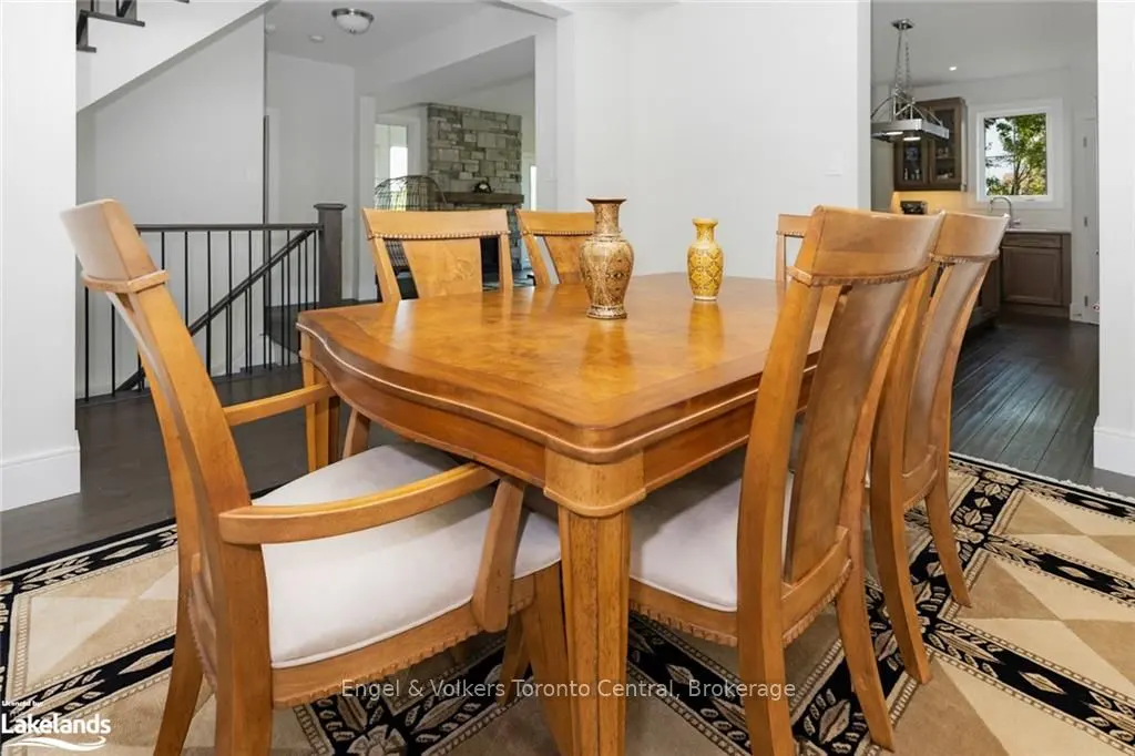 A dining room features a wooden table with six chairs, two vases, and a patterned rug. A staircase and kitchen are visible in the background.