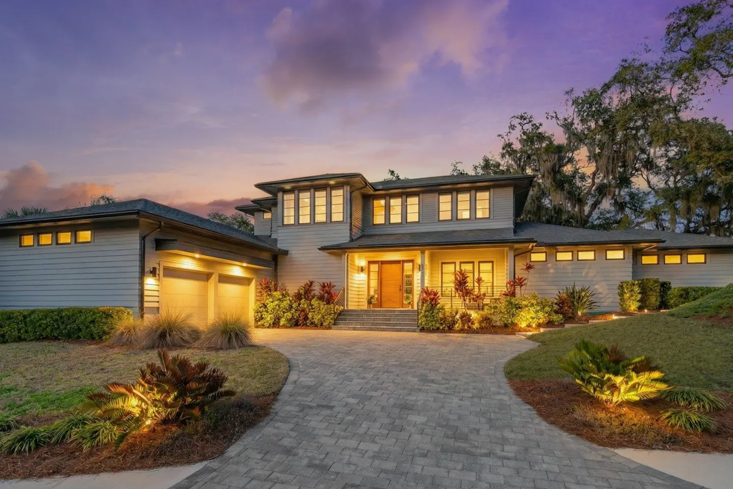 Two-story gray house with a brick driveway at dusk. The house has a two-car garage and a wooden front door.