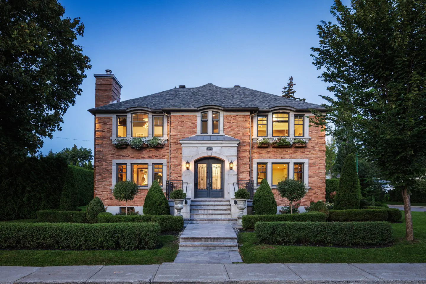 A two-story brick house with a gray roof, black trim, and manicured green hedges.