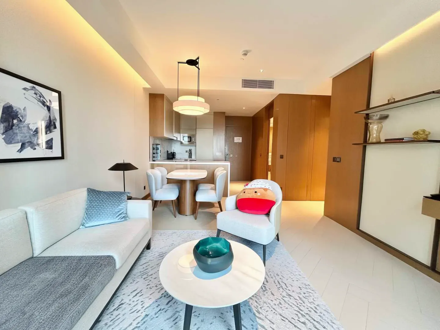 A modern apartment living room with a white sofa, round table, and open kitchen. The color palette is neutral with pops of blue and red.