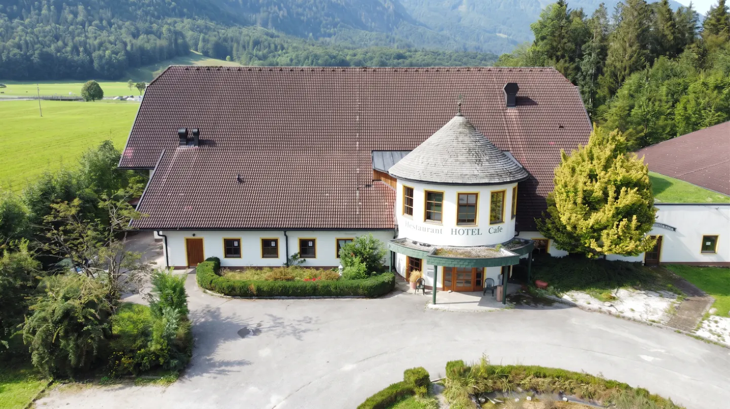 Aerial view of a white hotel with a brown roof, a turret, and green landscaping, set against a backdrop of green fields and mountains.