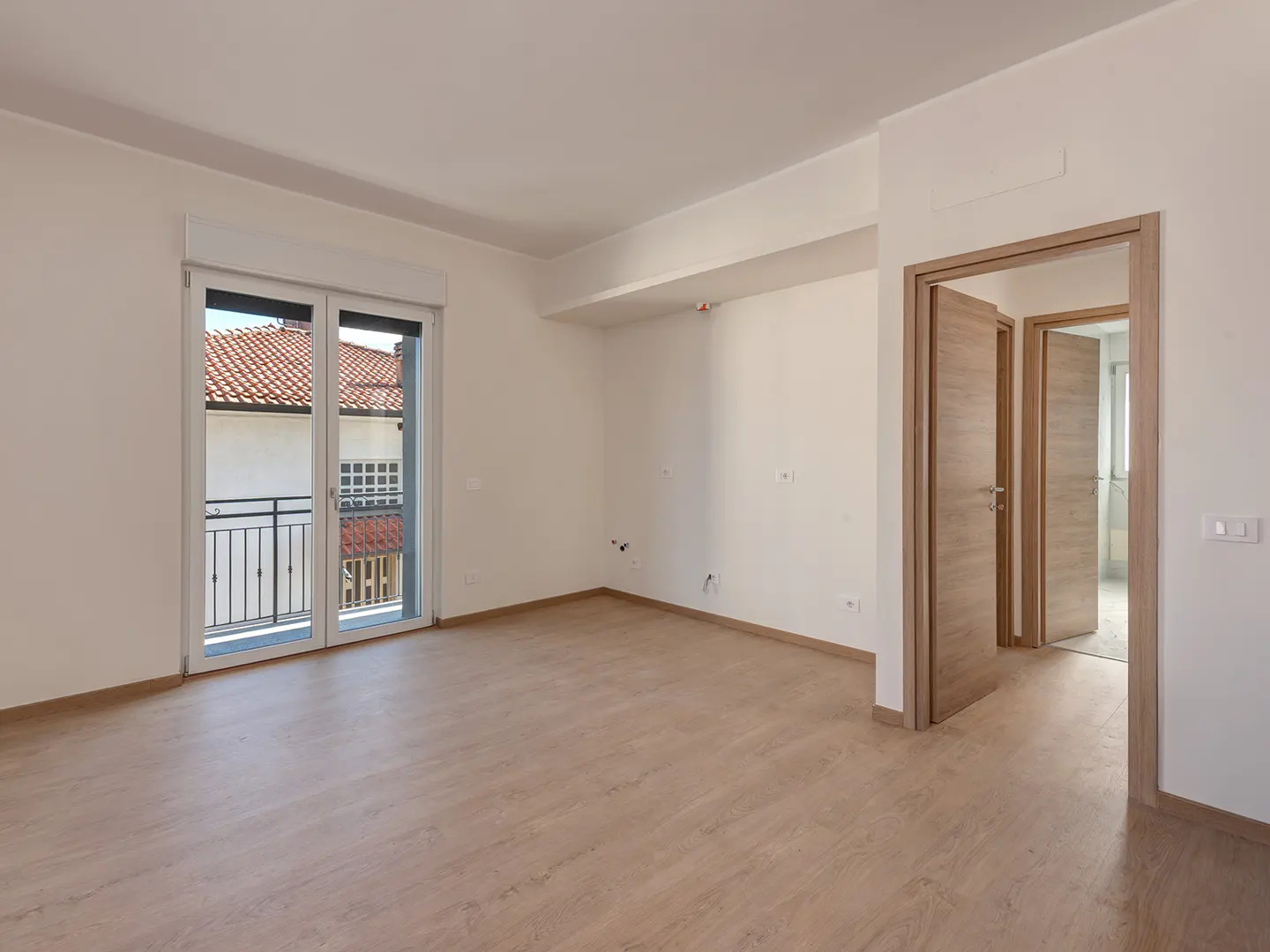 Bright, empty room with light wood floors, white walls, and a balcony door. Two open wood doors lead to other rooms.