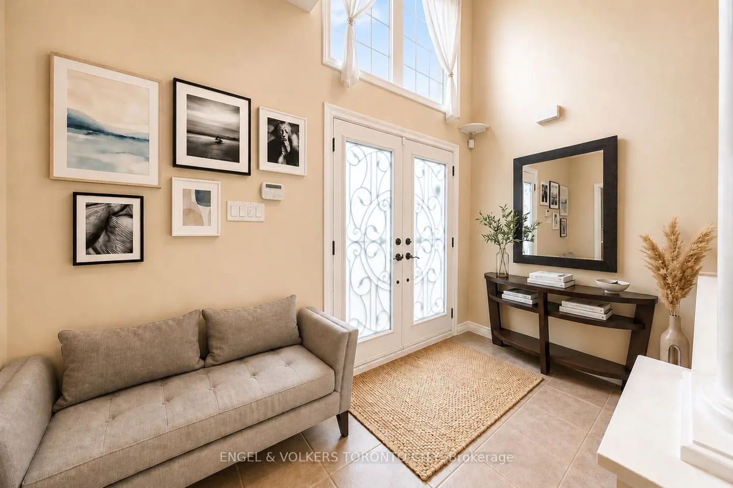 A beige foyer with a gray sofa, framed art, and a dark wood console table with a mirror above it. Double doors lead outside.