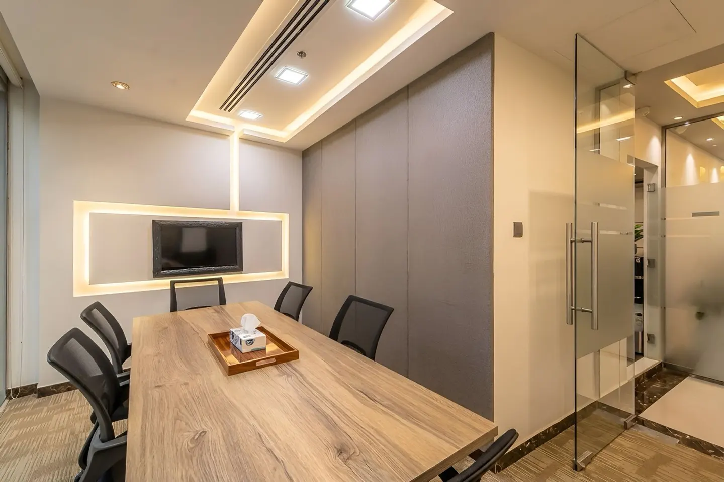 A modern conference room with a wood table, black chairs, and a wall-mounted TV. The room has a glass door and recessed lighting.