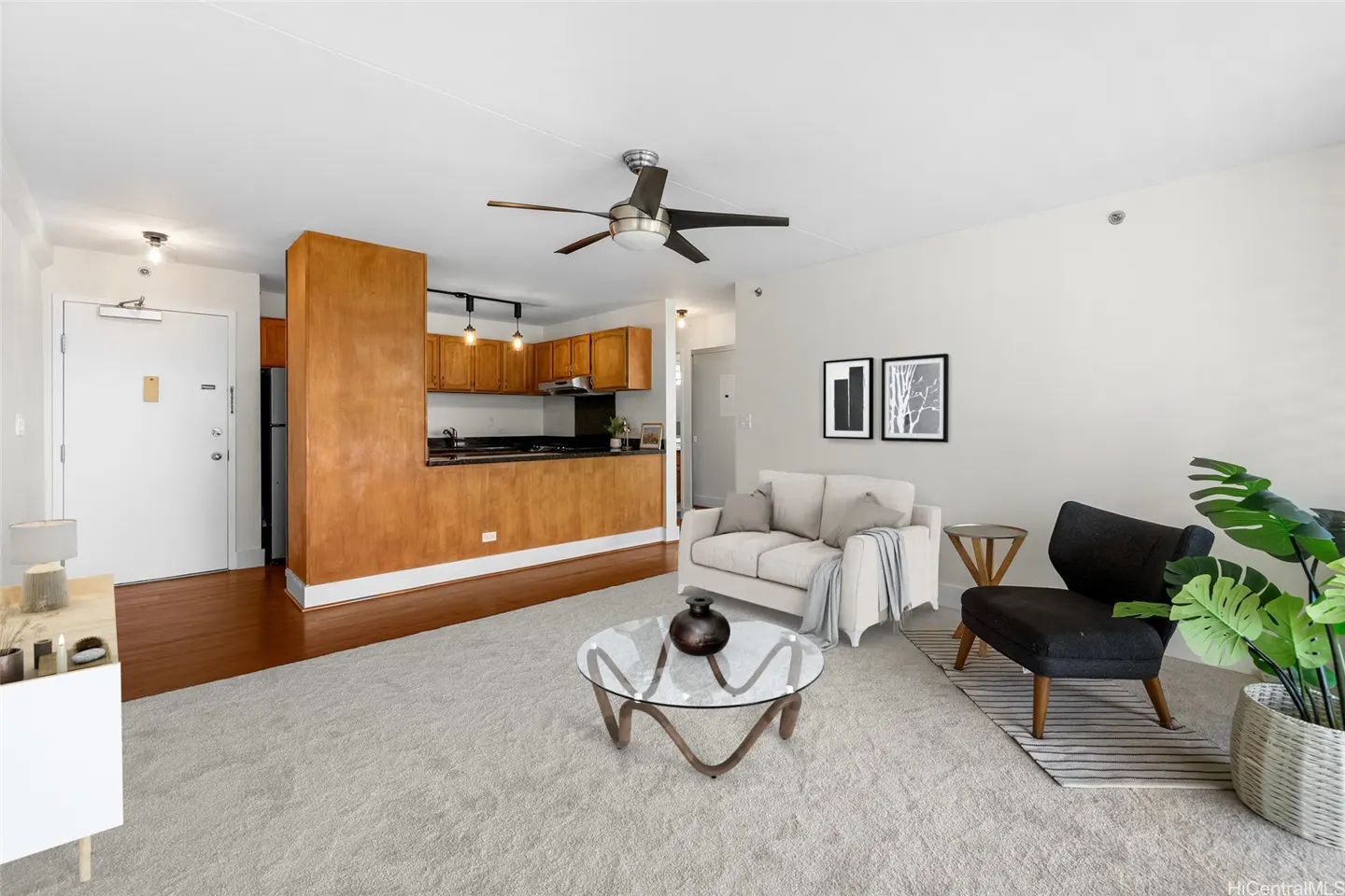 Open-concept living room with beige carpet, white sofa, black armchair, glass coffee table, and wood-paneled kitchen.