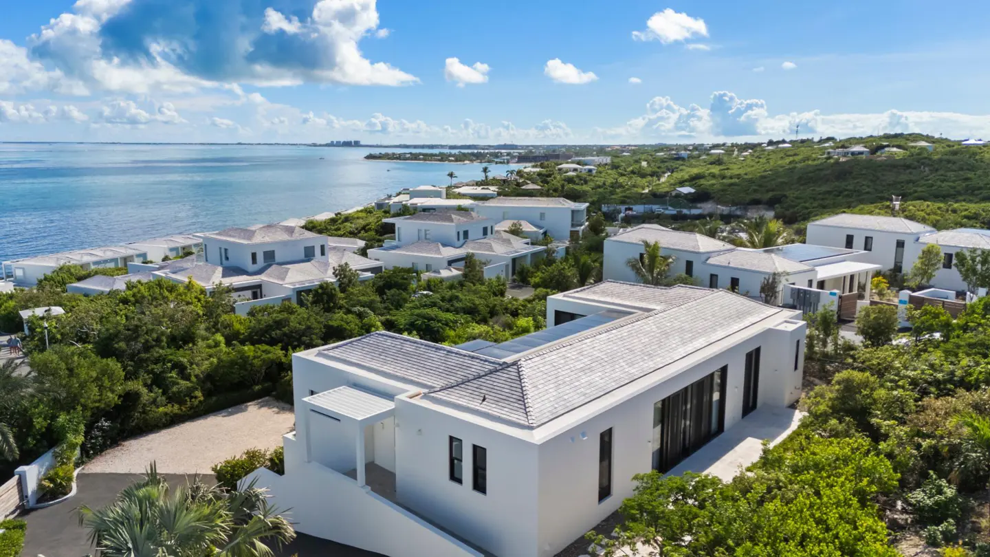 Aerial view of modern white houses with gray roofs surrounded by green trees near a blue ocean under a partly cloudy sky.