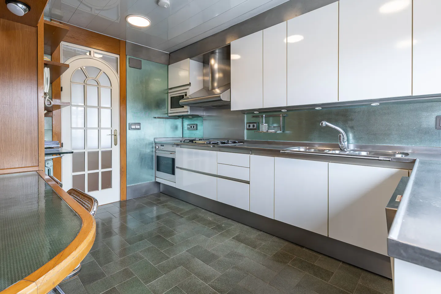 A modern kitchen with white cabinets, stainless steel appliances, and gray tile flooring. A glass-topped table with a wooden edge is in the foreground.