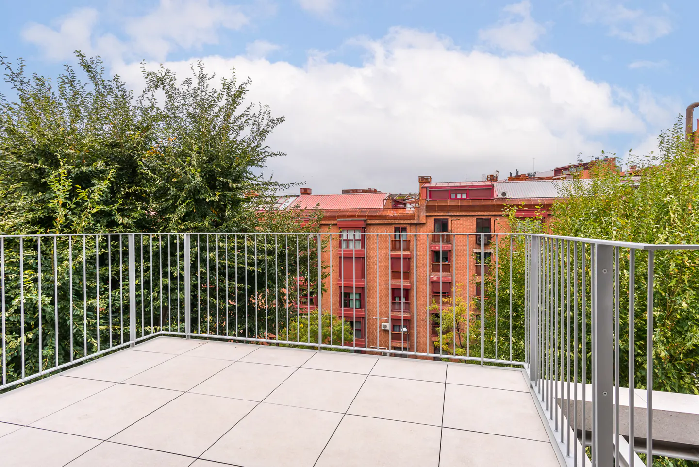 Balcony view with gray railings, light tile floor, trees, and a brick building with a red roof against a cloudy sky.