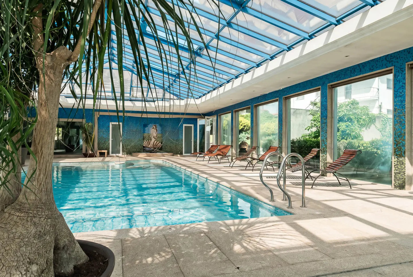 Indoor pool area with blue mosaic walls, lounge chairs, and a glass ceiling. A potted tree is in the foreground.
