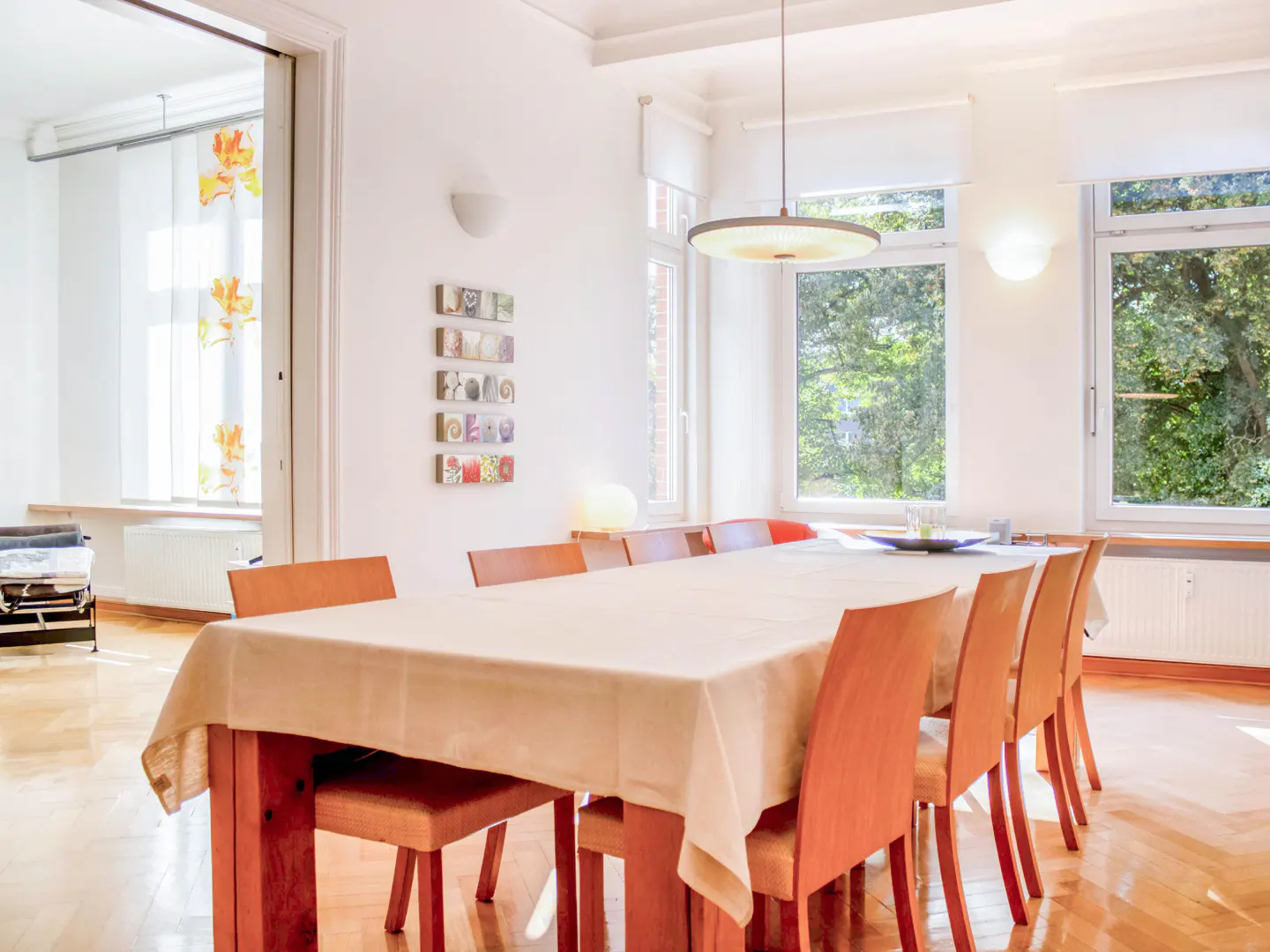 Bright dining room with a long table covered in a beige cloth, surrounded by orange chairs. Large windows overlook green trees.