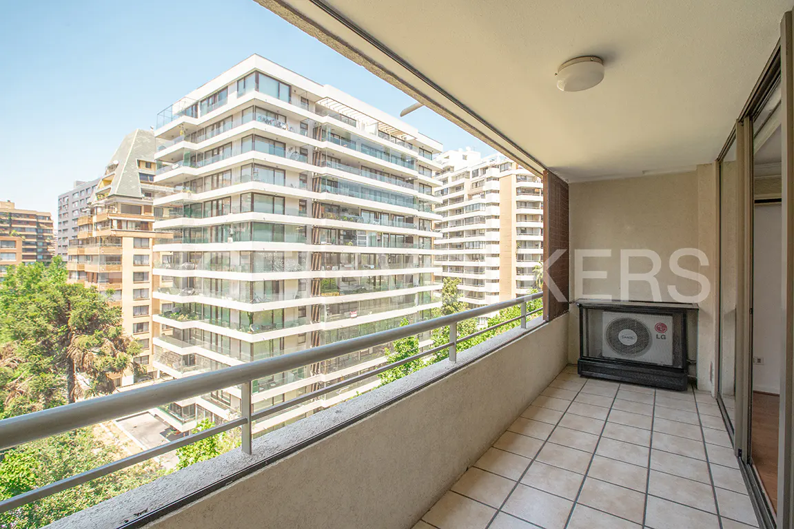 View from a balcony with beige tile flooring, a gray railing, and a view of tall buildings and trees. An air conditioning unit sits on the balcony.