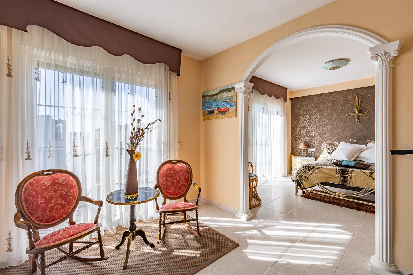 Living room with red rocking chairs, a table with a vase, and a view into a bedroom through an arched doorway.